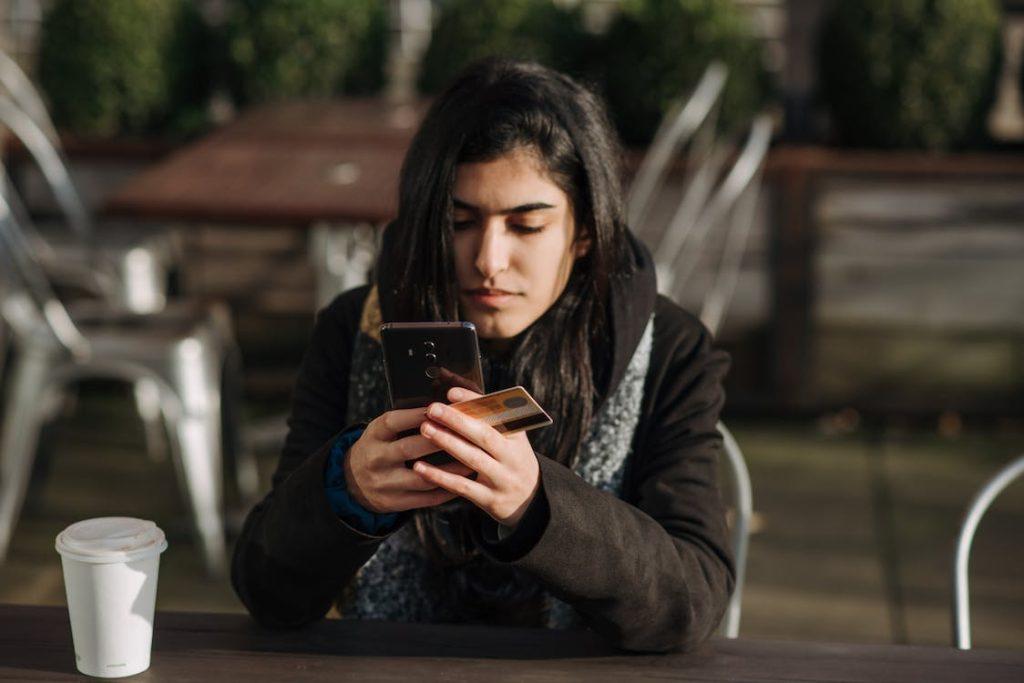 A person sat beside a coffee cup using their phone while holding a credit card