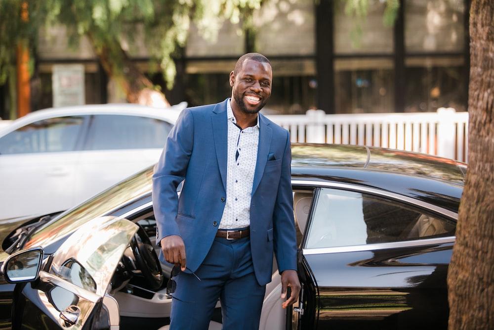 A man wearing a suit standing in front of a car holding sunglasses